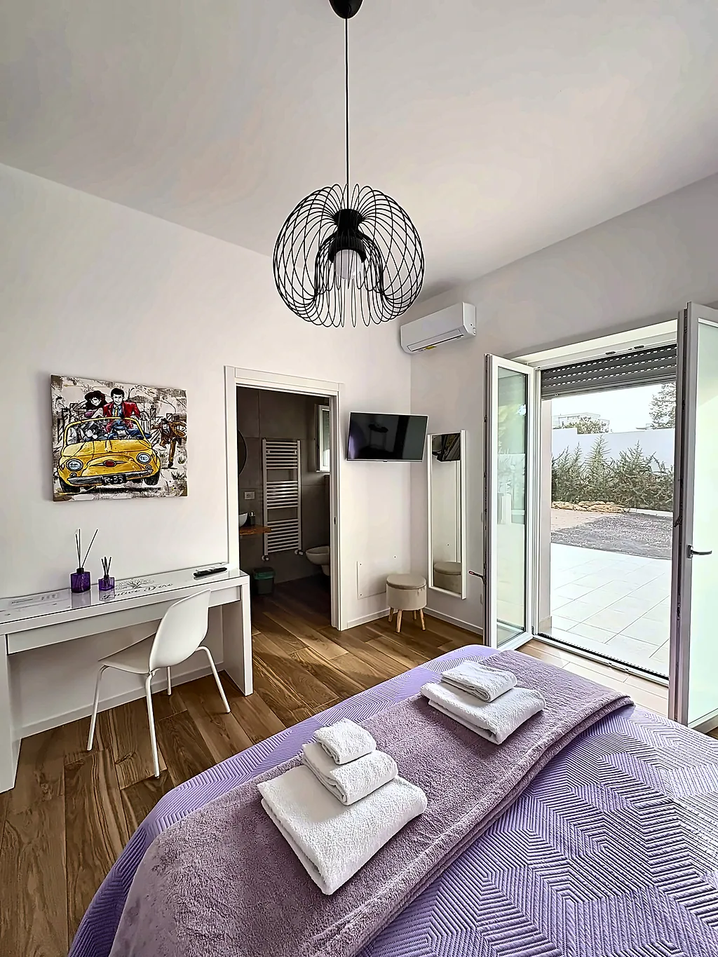 La scrivania, un quadro colorato e la portafinestra aperta, con il letto in primo piano