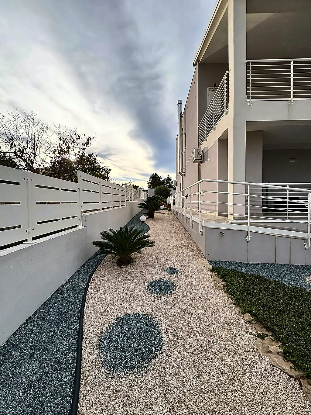 Il vialetto laterale in ghiaia chiara con pietre tonde, tra la recinzione bianca e la casa