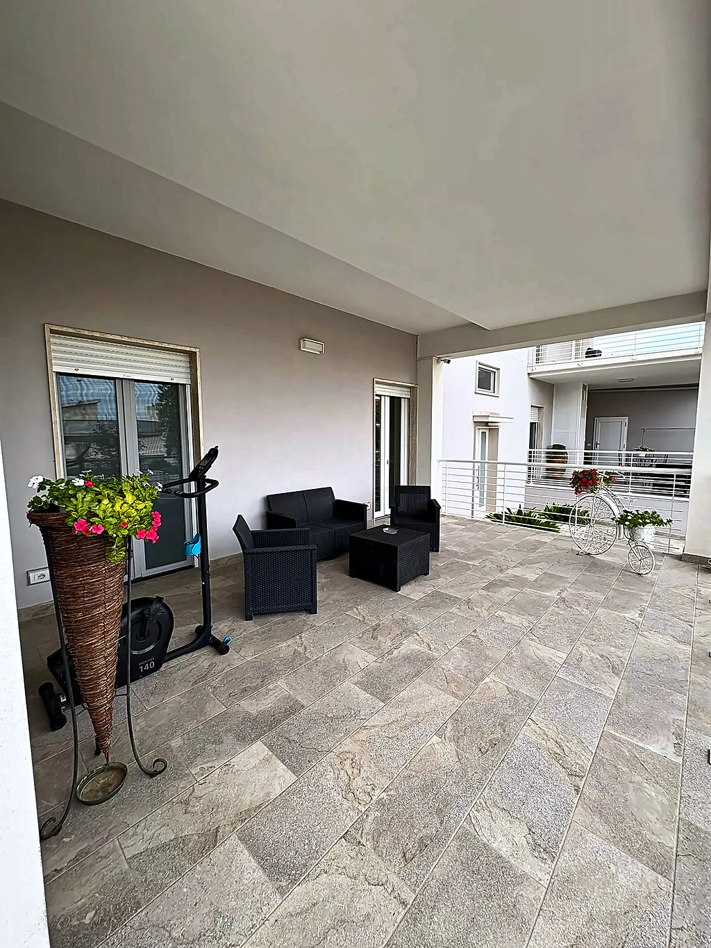 La veranda coperta con divanetti e tavolino da esterno, fiori e una cyclette