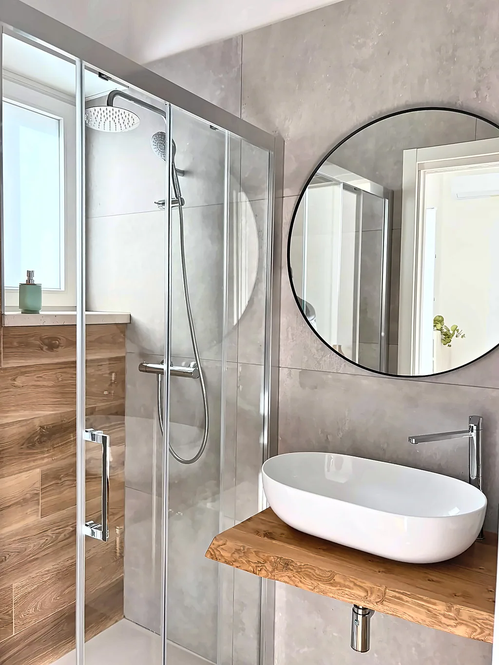 Il bagno della camera Ginkgo, con doccia in vetro, lavabo in appoggio su mensola in legno e specchio rotondo