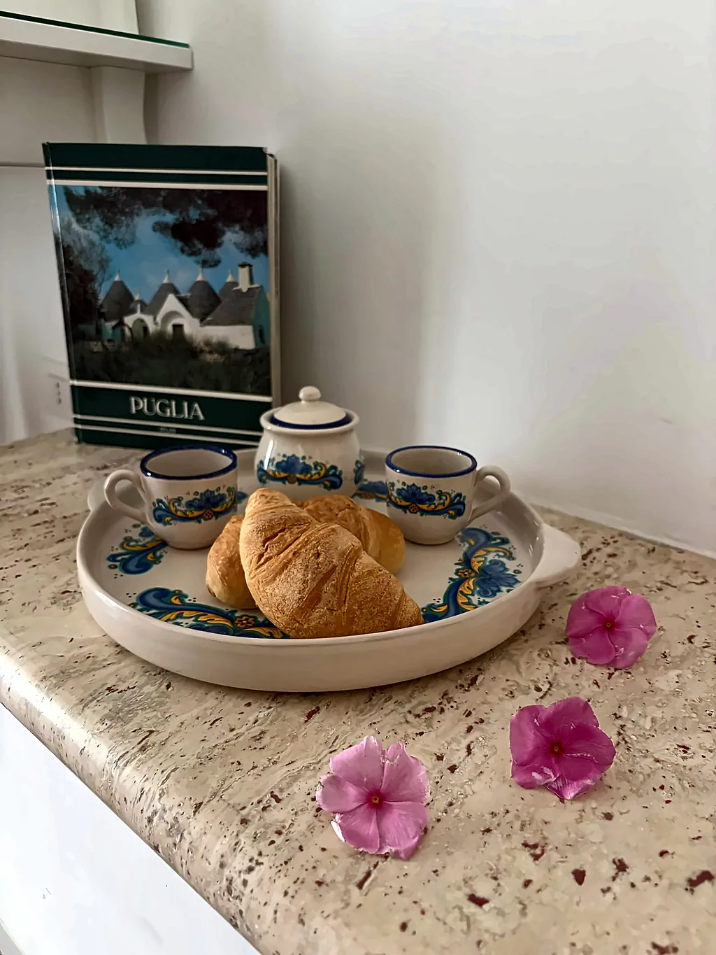Un vassoio in ceramica decorata con cornetti, tazzine e zuccheriera, accanto a un libro sulla Puglia e a qualche fiore